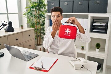 Canvas Print - Young hispanic doctor man holding first aid kit covering mouth with hand, shocked and afraid for mistake. surprised expression