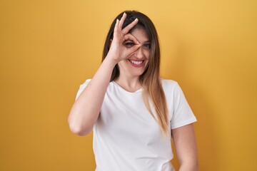 Wall Mural - Young brunette woman standing over yellow background doing ok gesture with hand smiling, eye looking through fingers with happy face.