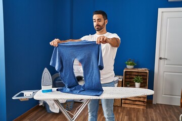 Sticker - Young hispanic man with beard ironing holding burned iron shirt at laundry room skeptic and nervous, frowning upset because of problem. negative person.