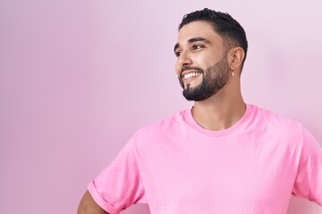 Wall Mural - Hispanic young man standing over pink background looking away to side with smile on face, natural expression. laughing confident.