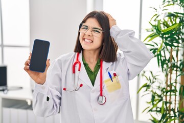 Canvas Print - Young hispanic doctor woman showing smartphone screen stressed and frustrated with hand on head, surprised and angry face