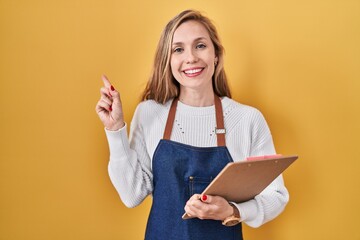 Sticker - Young blonde woman wearing professional waitress apron holding clipboard smiling happy pointing with hand and finger to the side