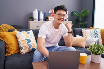 Wall Mural - Young asian man using laptop at home sitting on the sofa smiling with happy face looking and pointing to the side with thumb up.