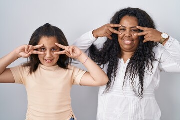 Poster - Mother and young daughter standing over white background doing peace symbol with fingers over face, smiling cheerful showing victory