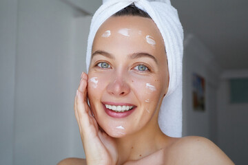 Young beautiful woman is applying  a cream, scrub beauty product on her face. Morning skin care routine.  Domestic daily skincare