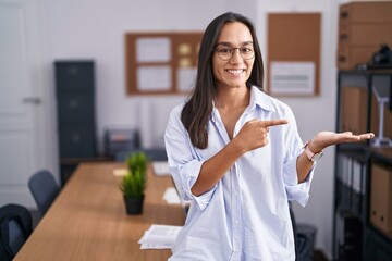 Canvas Print - Young hispanic woman at the office amazed and smiling to the camera while presenting with hand and pointing with finger.