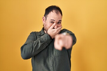 Canvas Print - Plus size hispanic man with beard standing over yellow background laughing at you, pointing finger to the camera with hand over mouth, shame expression