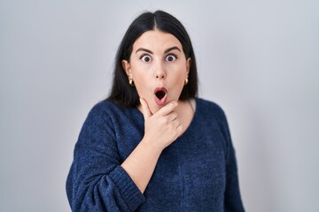 Canvas Print - Young brunette woman standing over isolated background looking fascinated with disbelief, surprise and amazed expression with hands on chin