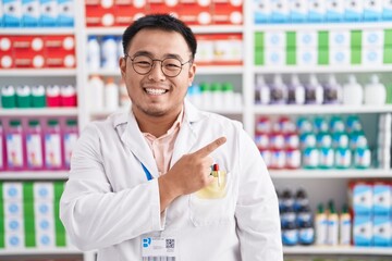 Poster - Chinese young man working at pharmacy drugstore cheerful with a smile on face pointing with hand and finger up to the side with happy and natural expression