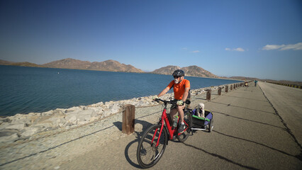 Elderly senior man biking on an e-bike on a trail pulling a trailer with a cute dog in it beside a lake with mountains in the distance.