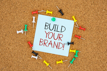 Poster - Build your brand text on light blue post-it paper pinned on bulletin cork board surrounding by multi color pins. This message can be used in business concept about building brand.