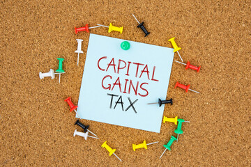 Poster - Capital gains tax text on light blue post-it paper pinned on bulletin cork board surrounding by multi color pins. This message can be used in business concept about capital gains tax.
