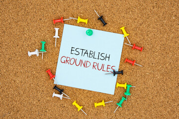 Poster - Establish ground rules text on light blue post-it paper pinned on bulletin cork board surrounding by multi color pins. This message can be used in business concept about establishing ground rules.