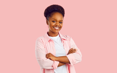 Wall Mural - Studio portrait of happy woman. Confident attractive young African American lady in casual pink shirt and white tee standing with her arms folded on pink background, looking at camera and smiling