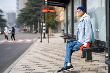 Young blond guy student waiting transport very long time in morning.Man on bus stop with coffee away. Blonde hair millennial man on bus stop in city. Modern urban transport for everyone