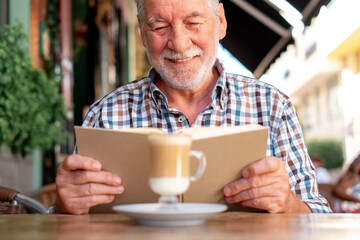 Sticker - Attractive senior man sitting outdoor at a cafe table reading a book while enjoying a coffee and milk drink - caucasian elderly man relaxed in retirement or vacation