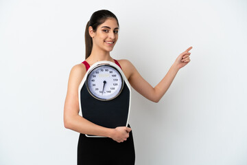 Wall Mural - Young caucasian woman isolated on white background with weighing machine and pointing side