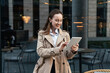 © muse studio - In a trench coat, a freelancer woman using a tablet is standing near the office. Leader surf the Internet, good mood.