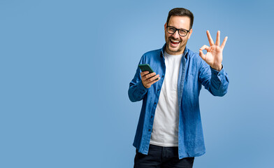 Portrait of satisfied businessman using smart phone and showing OK gesture against blue background