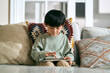 © imtmphoto - little asian boy sitting on family couch at home looking at and tapping the screen of digital tablet