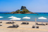 Fototapeta  - Deck chair and umbrella on beautiful Agios Stefanos Beach in front of paradise Island Kastri- historical ruins and paradise scenery at coast of island Kos, Greece