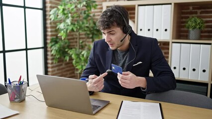 Wall Mural - Young hispanic man business worker wearing headset on a video call using credit card at office