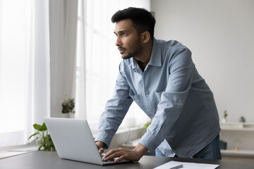 Wall Mural - Confident handsome young Indian business owner man working at laptop computer, standing at table, looking away in deep thoughts, thinking on creative ideas for project, making decision