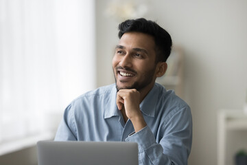 Happy young Indian business professional man thinking at home workplace table, looking at window away, smiling, laughing, working at laptop, planning successful project, career success