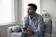 © fizkes - Serious thoughtful young adult Indian guy holding digital gadget, sitting on home couch, looking away lost in deep thoughts. Freelancer man, entrepreneur using Internet technology for work, thinking