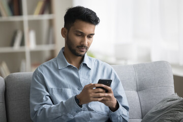 Serious handsome young Indian man using gadget for Internet communication at home, holding smartphone, working on business project with online technology, job application