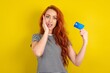 Astonished positive young beautiful red haired woman wearing striped shirt over yellow studio background arm face hold bank plastic card impressed functions