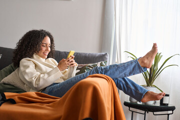 smiling relaxed young latin woman sitting on couch using cell phone at home. happy barefoot girl hol