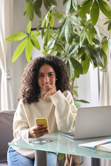 Wall Mural - Young happy smiling pretty curly latin woman holding smartphone using cell device modern technology, looking at camera with mobile phone in hand sitting at table a home, vertical shot.