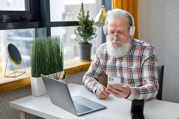 Handsome senior caucasian man sitting alone, using modern laptop and headset, bearded male having video call on smartphone, attending video conference or webinar, use modern technologies