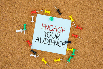 Poster - Engage your audience text on light blue post-it paper pinned on bulletin cork board surrounding by multi color pins. This message can be used in business concept about engage your audience.