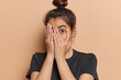 © WHstudio Leushin N - Scared Latin woman with hair bun peeks through her fingers fearfully hiding her face unable to bear sight of something terrible witnessing something truly terrible dressed in casual t shirt isolated