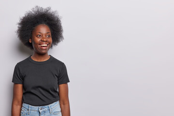 Horizontal shot of pretty dark skinned woman wtih curly bushy hair smiles broadly being very happy dressed in casual black tshirt and jeans isolated over white background copy space for your promotion