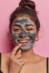 Self care beauty concept. Positive dark haired woman winks eye and smiles broadly applies charcoal facial mask for skin treatment stands indoor against pink background has refreshed radiant complexion