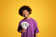 © Prostock-studio - Cheerful black adult curly man in purple t-shirt hold many dollar cash, isolated on orange background