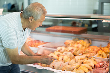 Mature man buyer of meat department of grocery store, looking at hen in refrigerator window. Elderly male customer points with finger and orders whole chicken fresh poultry