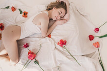 Young Redhead Woman Sleep In Bed With Red Tulips. Minimalist Light Pure Shoot. Concept Of Female Health, Menstrual Cycle And Ovulation. Allegorical Depiction Of Defloration. Top View