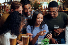 Happy Young Multiracial Group Of Friends In Casual Clothing Using Mobile Phone At Bar