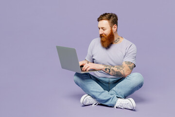 Full body young redhead bearded man wear violet t-shirt casual clothes sitting hold use work on laptop pc computer isolated on plain pastel light purple background studio portrait. Lifestyle concept.