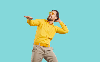 Happy man in modern stylish outfit dancing and having fun in fashion studio. Funny carefree guy wearing yellow shirt, beige pants, and cool trendy sunglasses dancing isolated on turquoise background