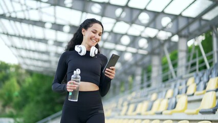 Young happy sportswoman using a smartphone while walking around the urban city stadium. Smiling female athlete typing a message, chatting online, browsing social media, watching reading notifications