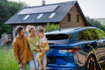 Family with little girl standing in front of their house with solar panels on the roof, having electric car.