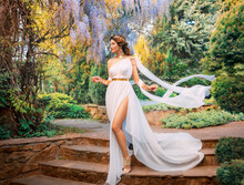 Fantasy Woman Greek Goddess Walks In Spring Garden Purple Flowers Green Tree Nature Day Sun Light. Sexy Girl Queen Antique Style White Silk Dress Fly, Gold Diadem On Red Hair, Lady Going Down Stairs