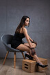 © Budjak Studio - Classic dark studio portrait of a young brunette woman in black clothes who is sitting on a chair against a concrete wall.