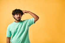 The Emotion Of Doubt, Reflection And Confusion. Of A Handsome Young Man In A Turquoise T-shirt On A Yellow Background, Scratching His Head And Looking Very Pensively To The Side. Copy Space.