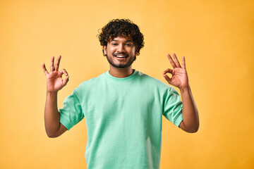 Handsome Indian man showing ok gesture, looking at camera and smiling while standing against yellow background.